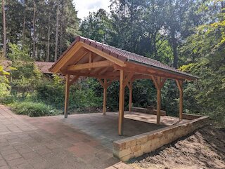 Freistehender Holzcarport mit Walmdach und offenen Holzbögen auf einem Waldgrundstück