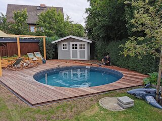 Holzterrasse mit rundem Pool, Sitzecke und Gartenhaus im begrünten Garten